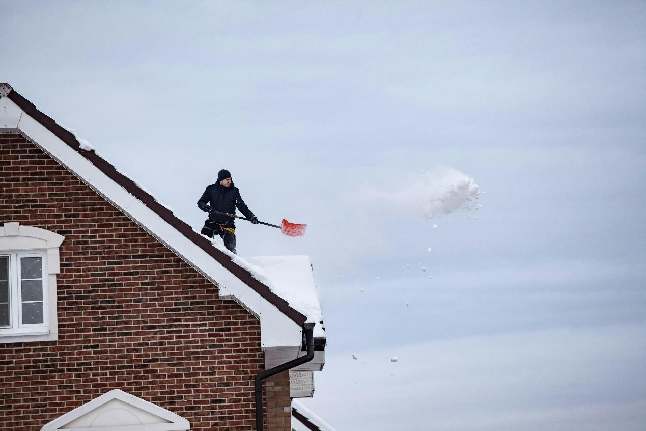 A picture of a man cleaning snow off a roof while wearing a black jacket and beanie.